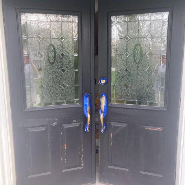 The same double front doors in worn charcoal paint, chipped along the edges, handles masked off for prep