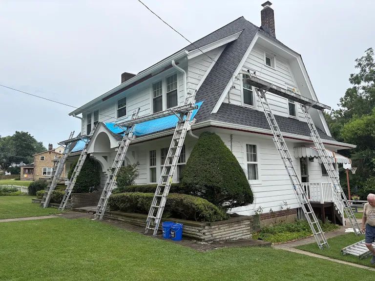 Large white gambrel-roofed house with ladders and staging planks set across the front and blue tarps over the porch roof