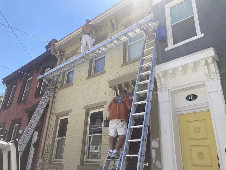 Two Caridi painters on ladders and a ladder-jack plank, prepping the cornice and upper facade of a cream brick rowhouse