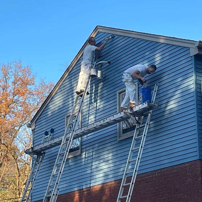 Two painters working from ladder jacks across the blue lap siding of a tall gable end