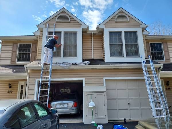 Caridi painter on a ladder cutting in the second-floor window trim of a tan townhouse, with a drop cloth spread over the garage roof below